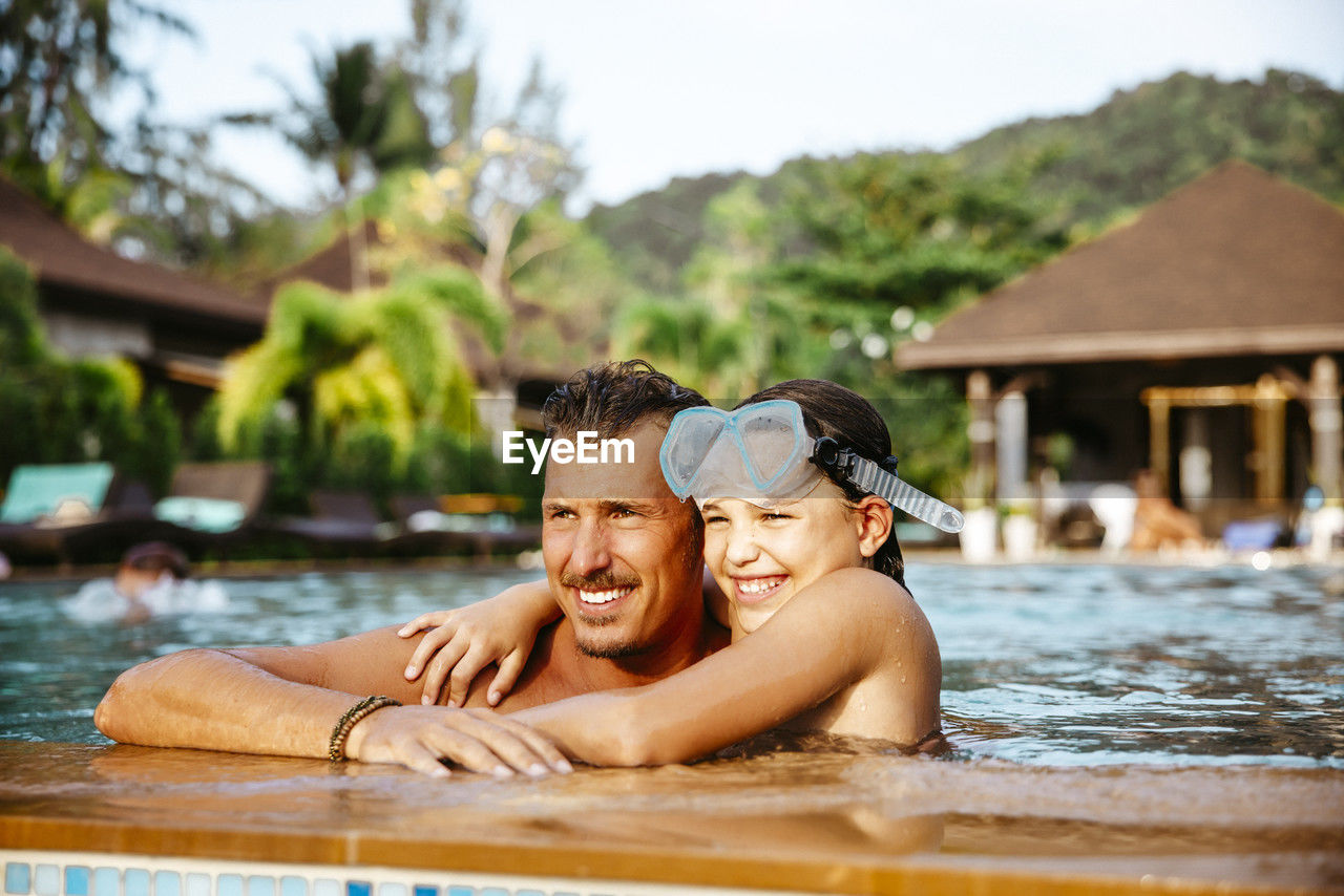 Smiling girl with arm around father in swimming pool at resort during vacation