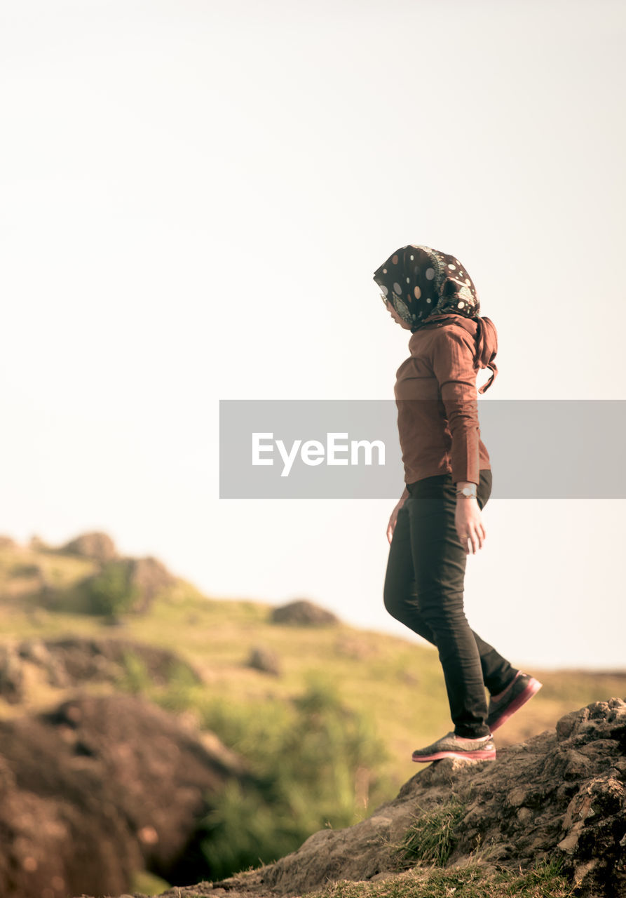 Side view of young woman standing on rock against clear sky