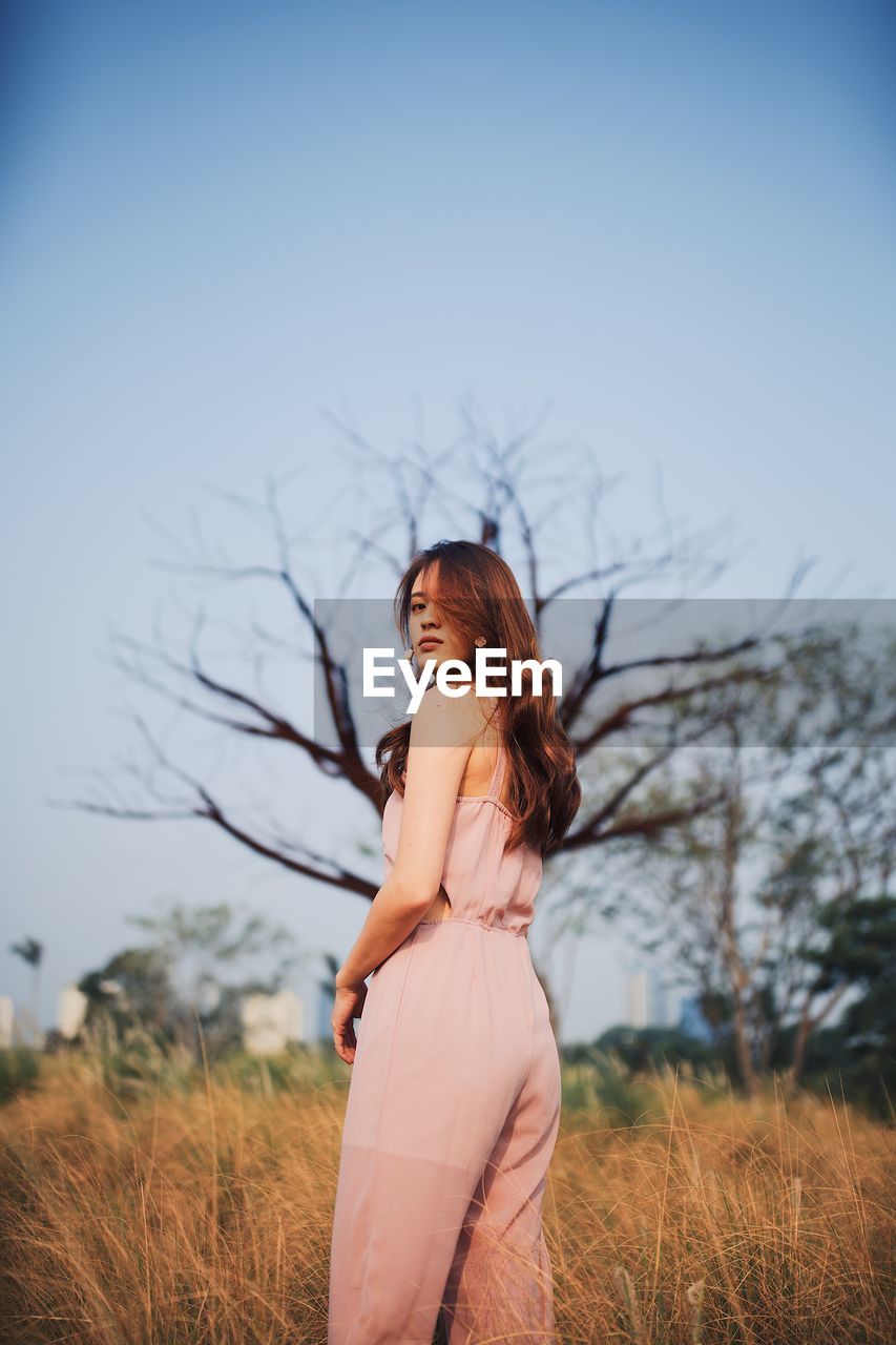 Portrait of young woman standing on grassy land against clear sky