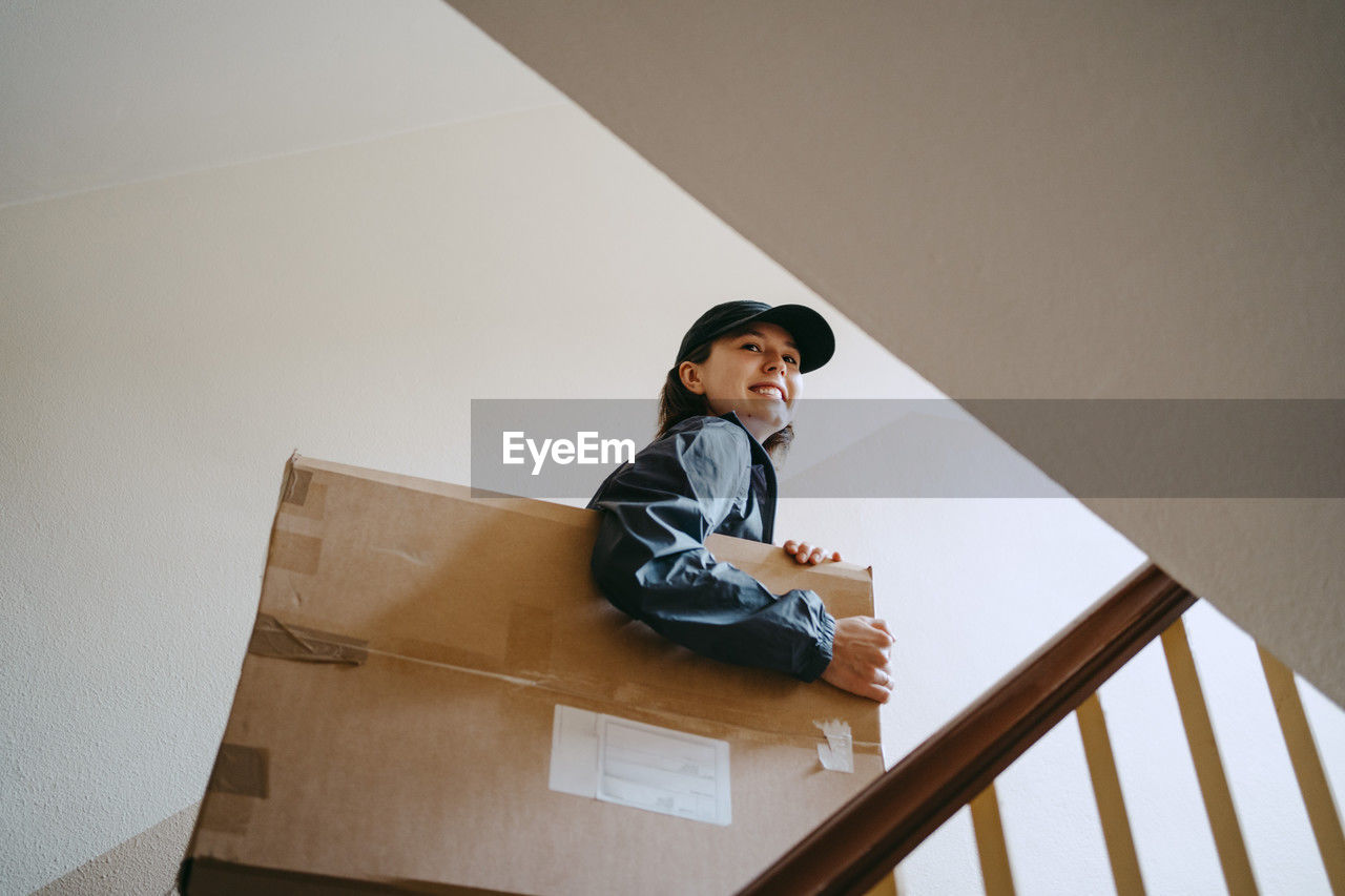 Low angle view of smiling female delivery person holding cardboard box while moving up on staircase in building