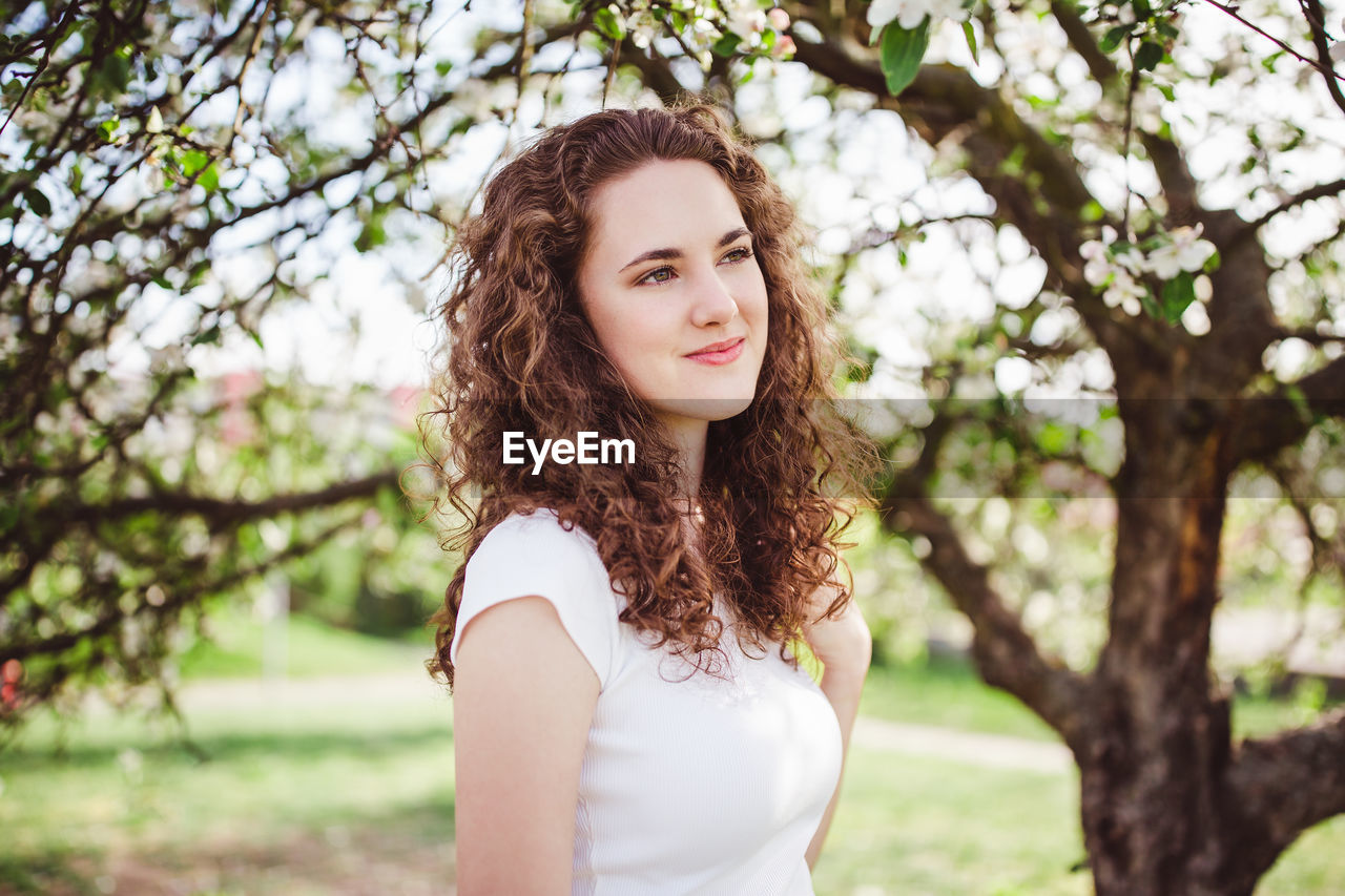 Happy curly hair young woman in a white t-shirt under the blooming apple tree. smiling.