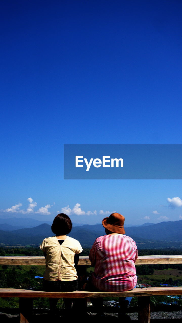 Rear view of friends looking at landscape while sitting on bench against blue sky