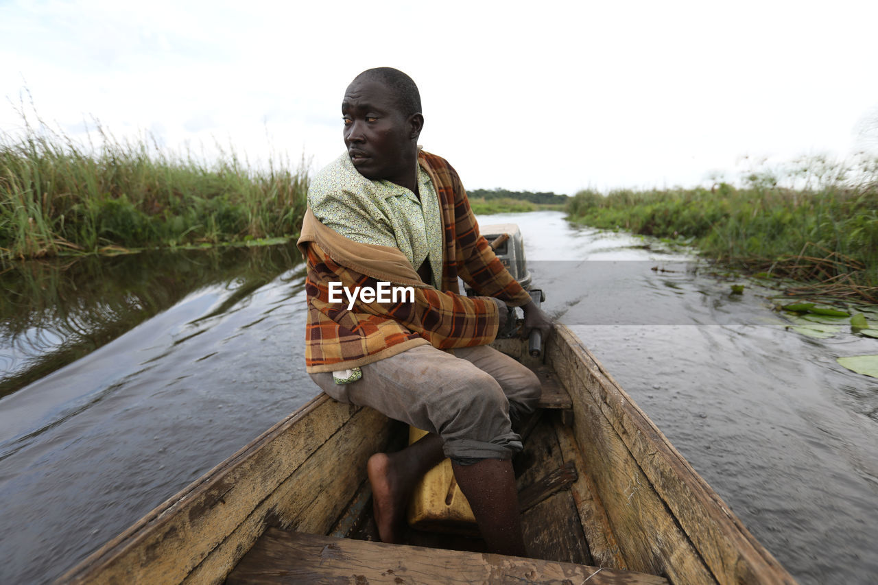 PORTRAIT OF MAN SITTING ON WOOD AGAINST LAKE