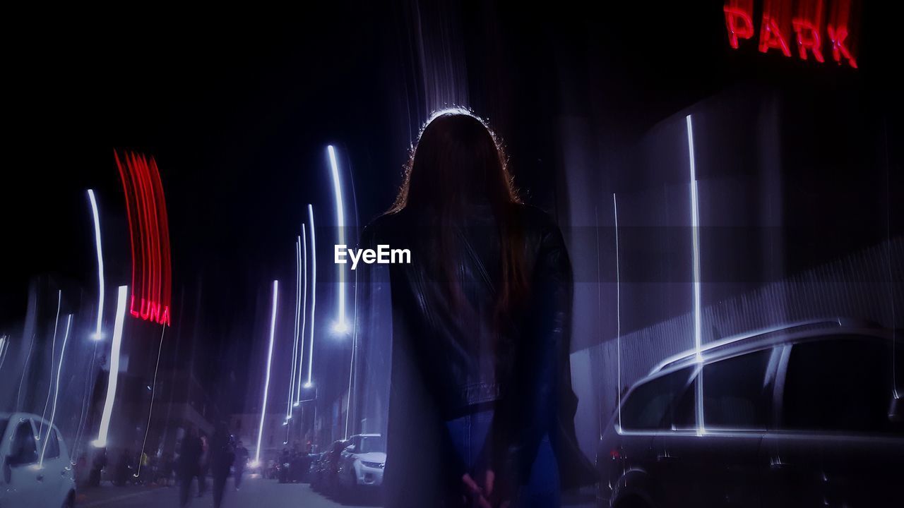 Rear view of woman standing on road in illuminated city at night