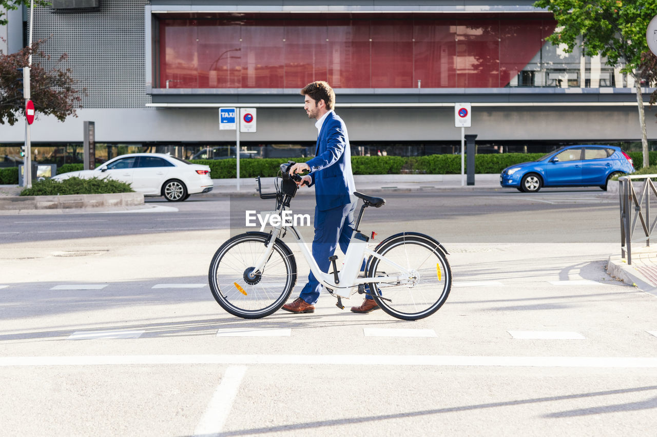 Side view of trendy unshaven male office worker in formal wear strolling on street with electric bicycle