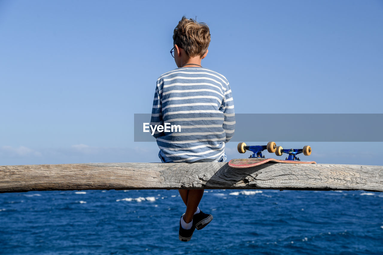 Rear view of boy sitting on wooden log over sea against clear blue sky