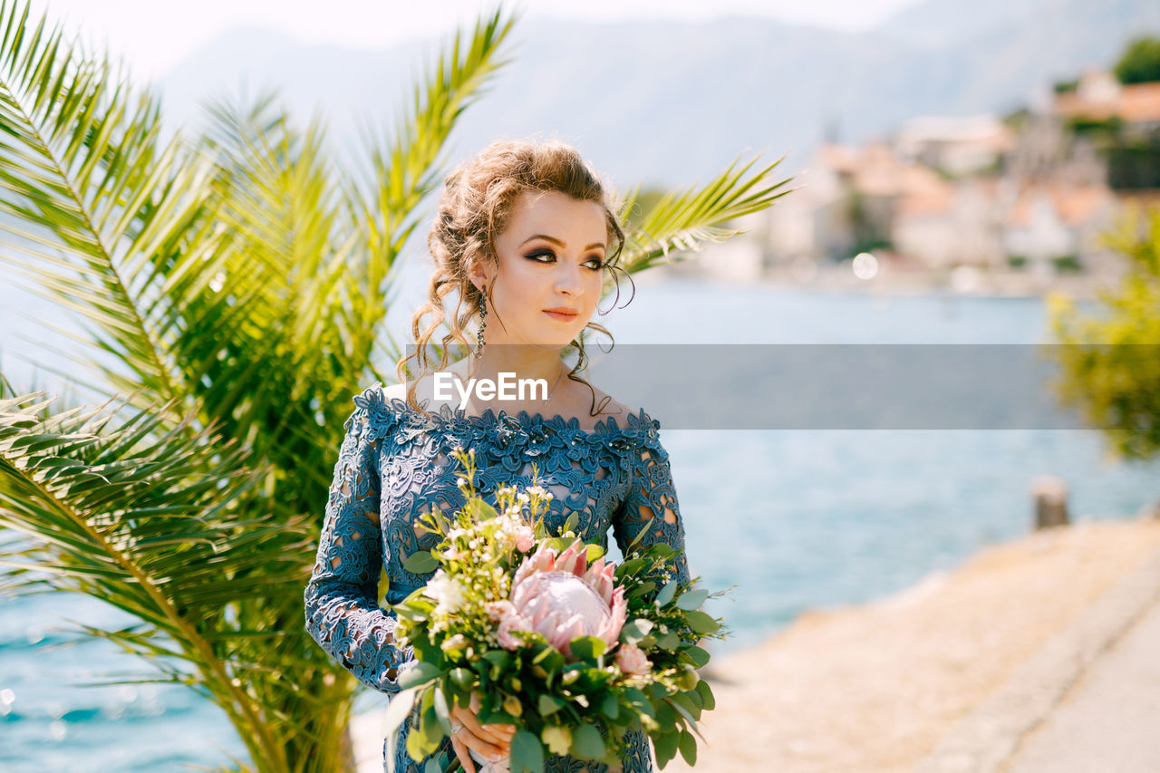 Bride holding bouquet while standing against sea