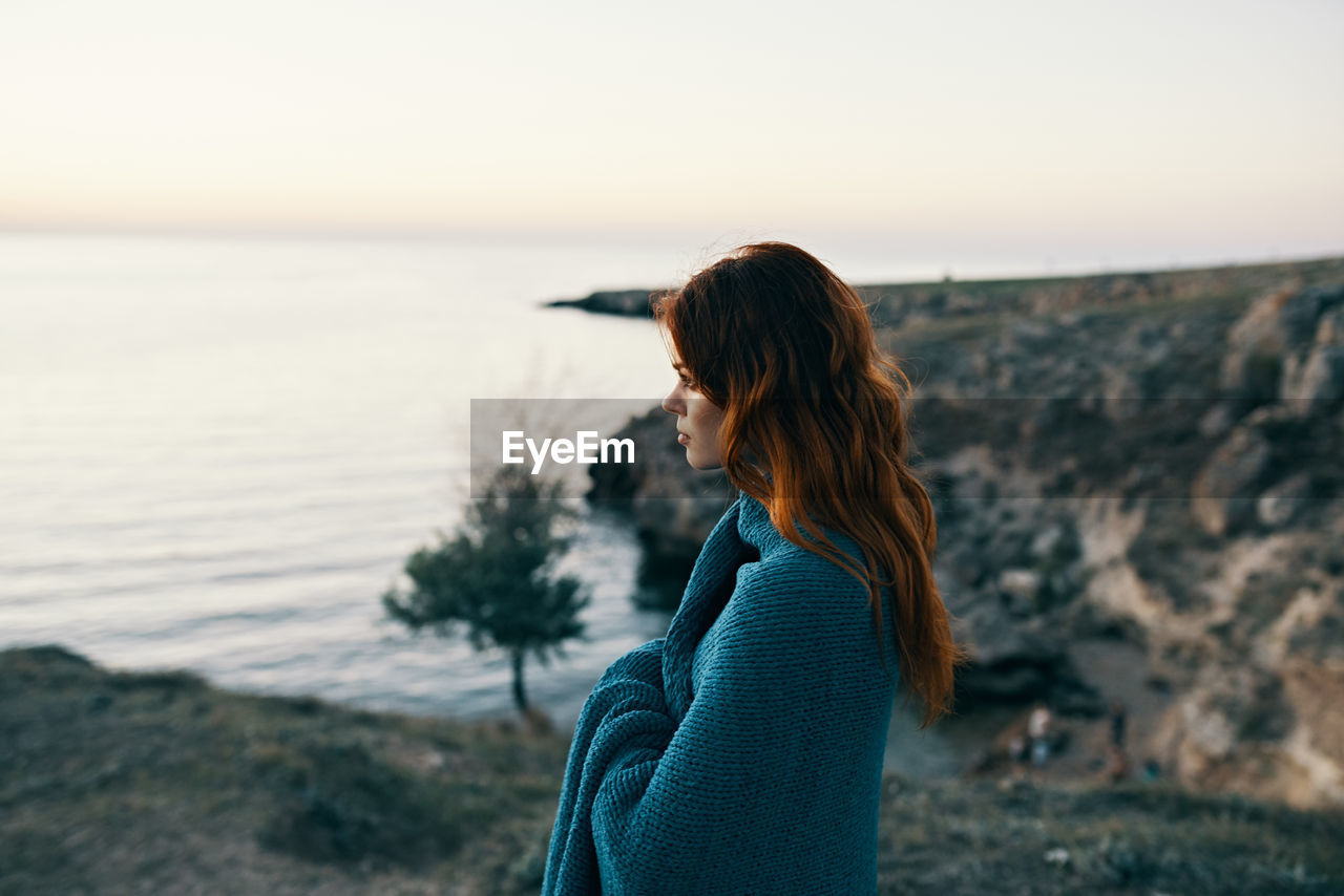 SIDE VIEW OF WOMAN STANDING AT SHORE AGAINST SKY