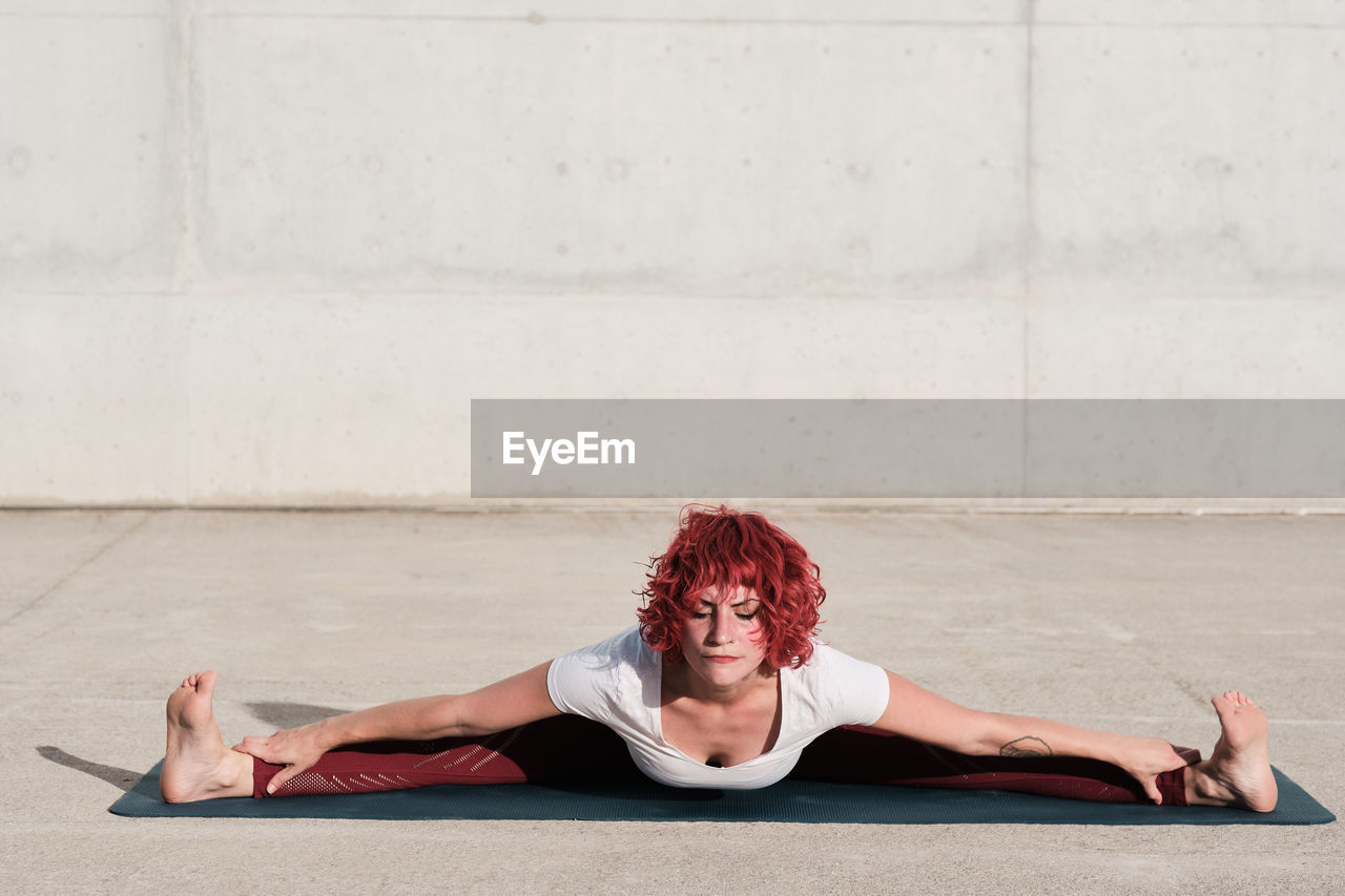 From above of barefooted woman with with closed eyes in sportswear doing yoga in wide angle seated forward bend pose on mat training alone on street against concrete wall