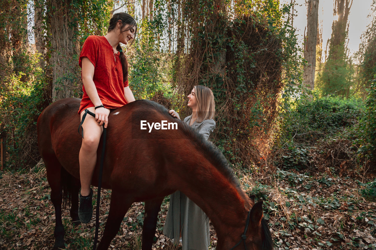 Young woman sitting on horse talking with friend in forest