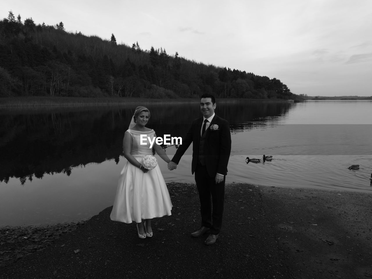 Portrait of wedding couple holding hands while standing at lakeshore against sky