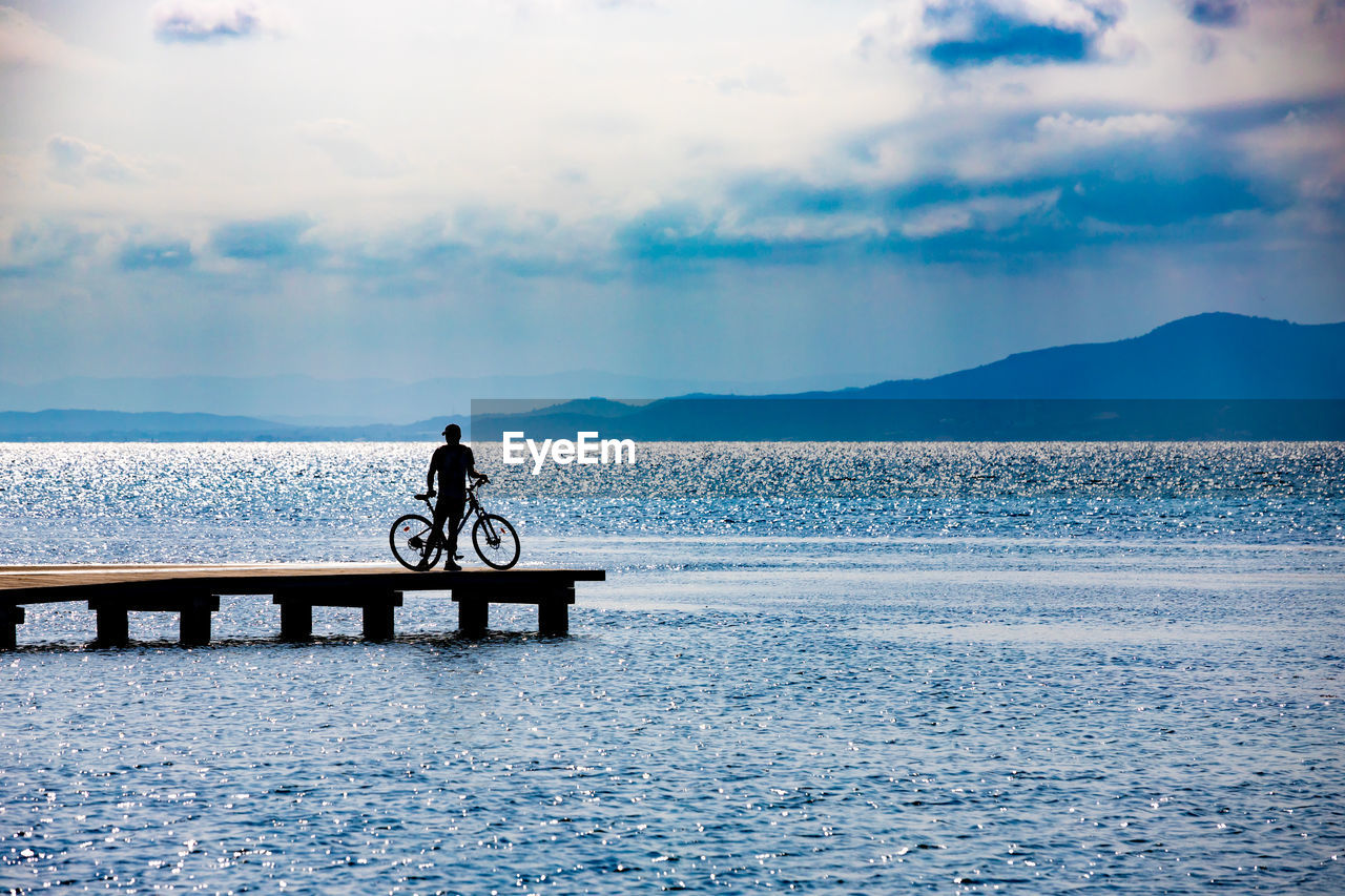 Silhouette man standing with bicycle on pier over sea against cloudy sky