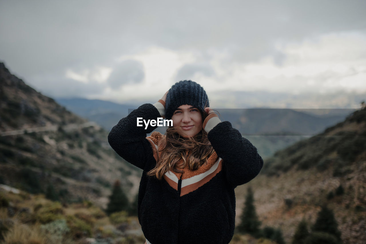 Smiling young female putting on warm hat and looking at camera while standing on rough highlands on cloudy gloomy day