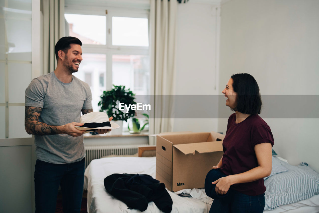 Happy man with hat talking to woman folding clothes by bed at home