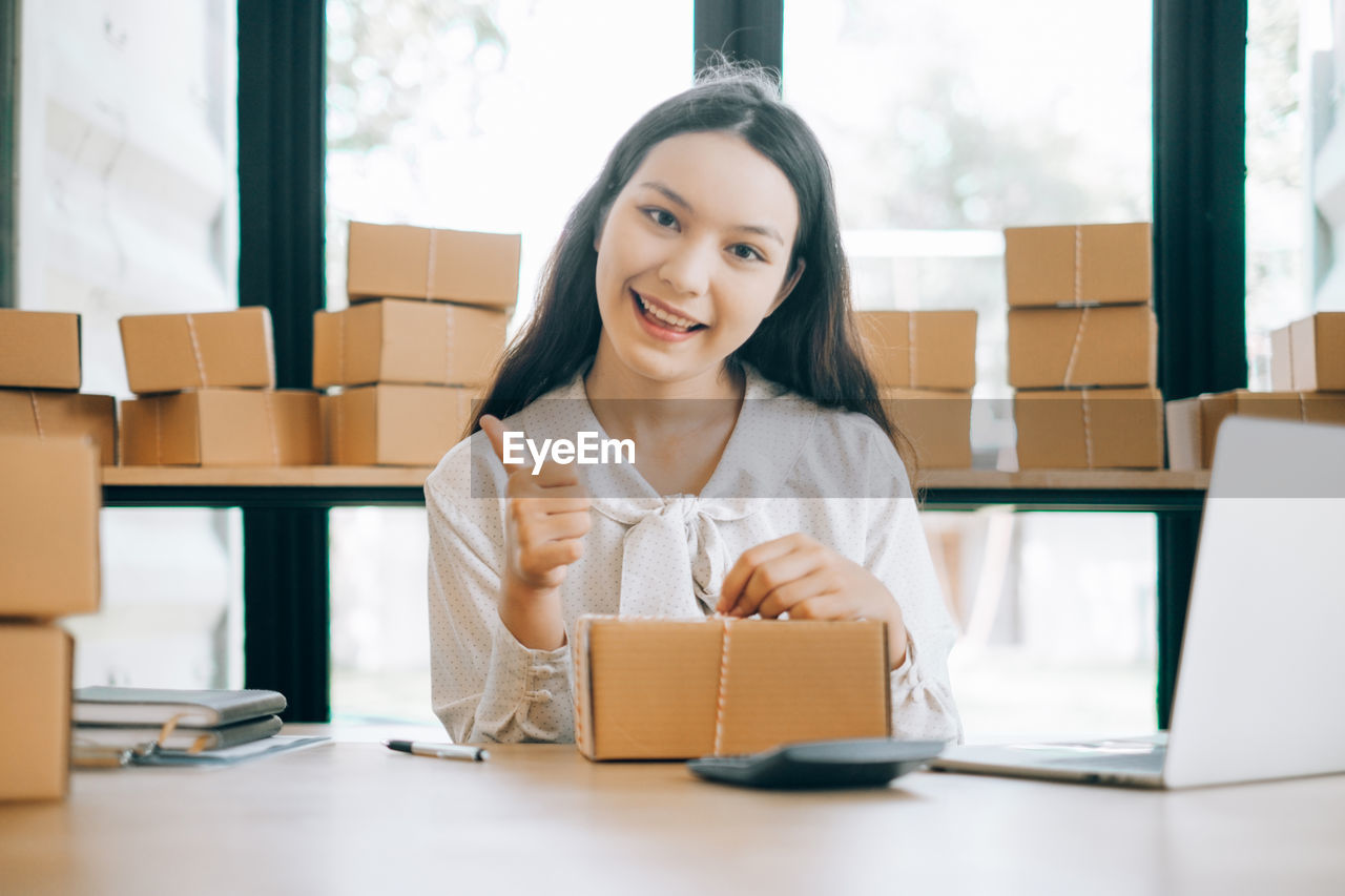 Portrait of smiling young woman showing thumbs up sign while packing cardboard box at office