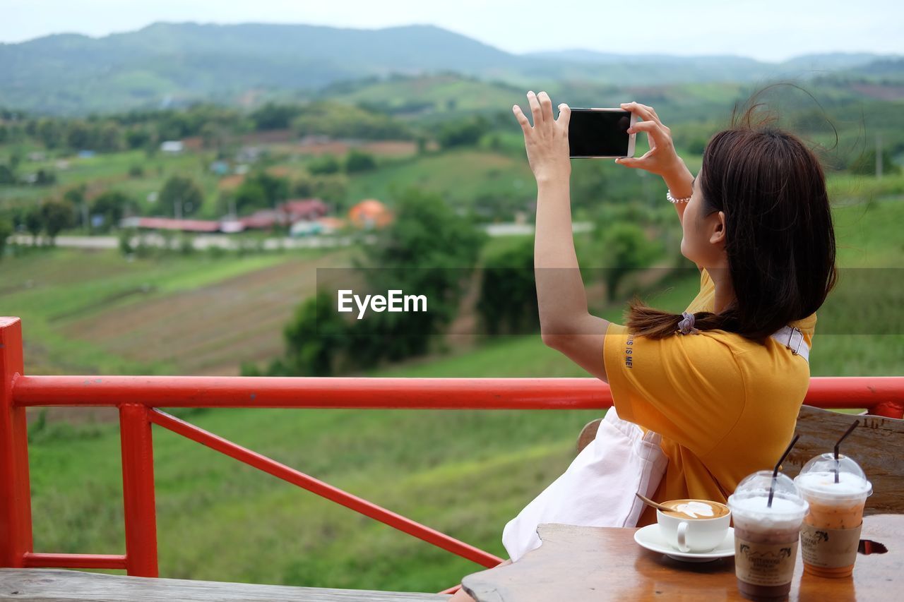 REAR VIEW OF WOMAN STANDING ON MOBILE PHONE AGAINST MOUNTAIN