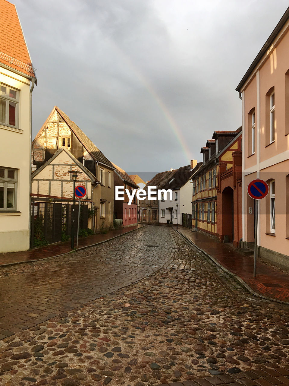 An empty cobbled street of a european city with old houses and a rainbow in the sky.
