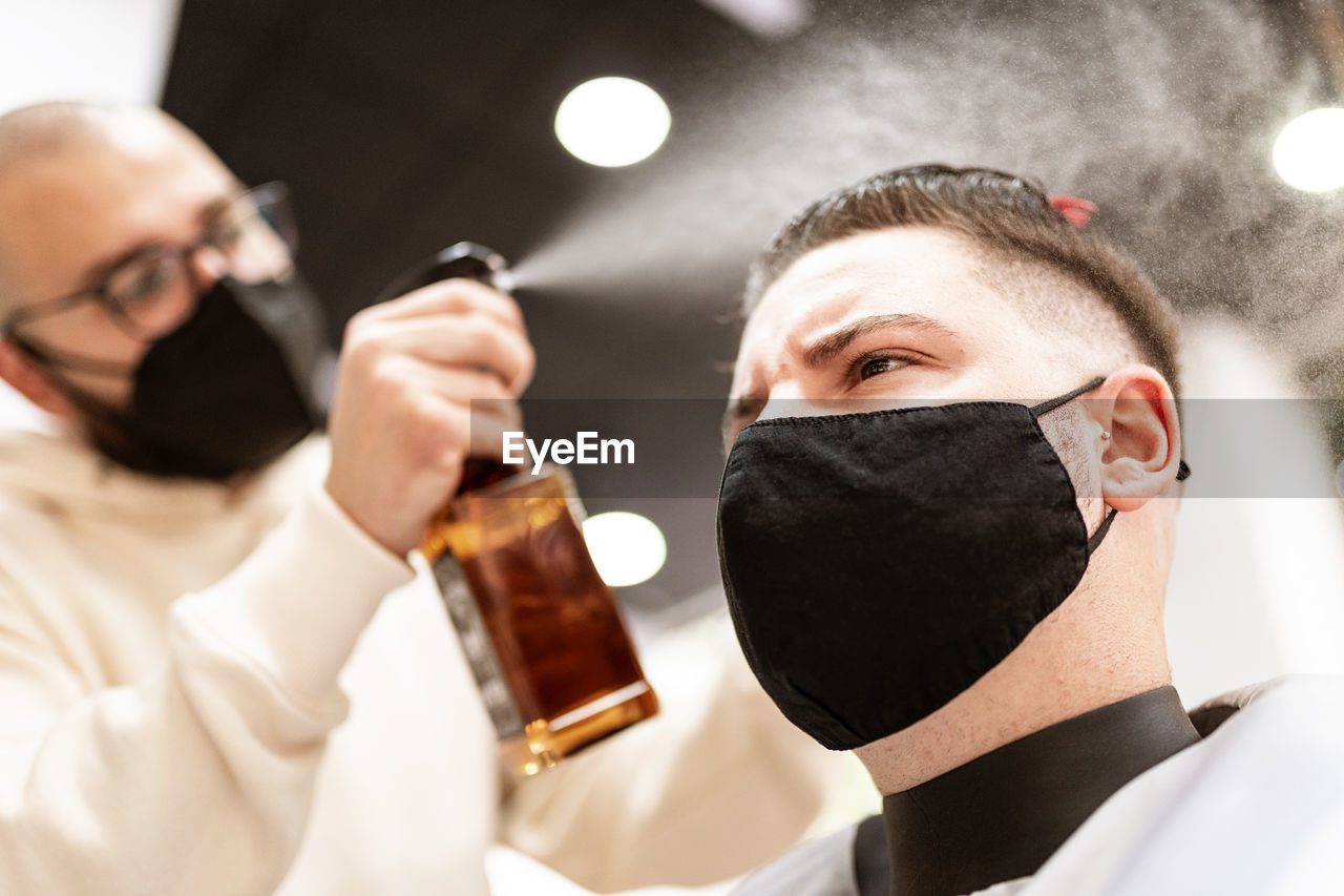 Male stylist in textile mask spraying haircut of client with liquid from bottle in barbershop