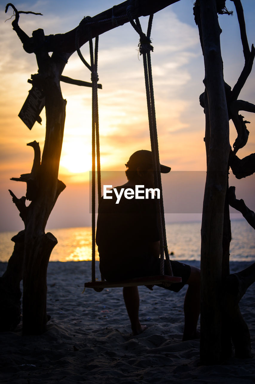 Silhouette man sitting on swing beach against sky during sunset