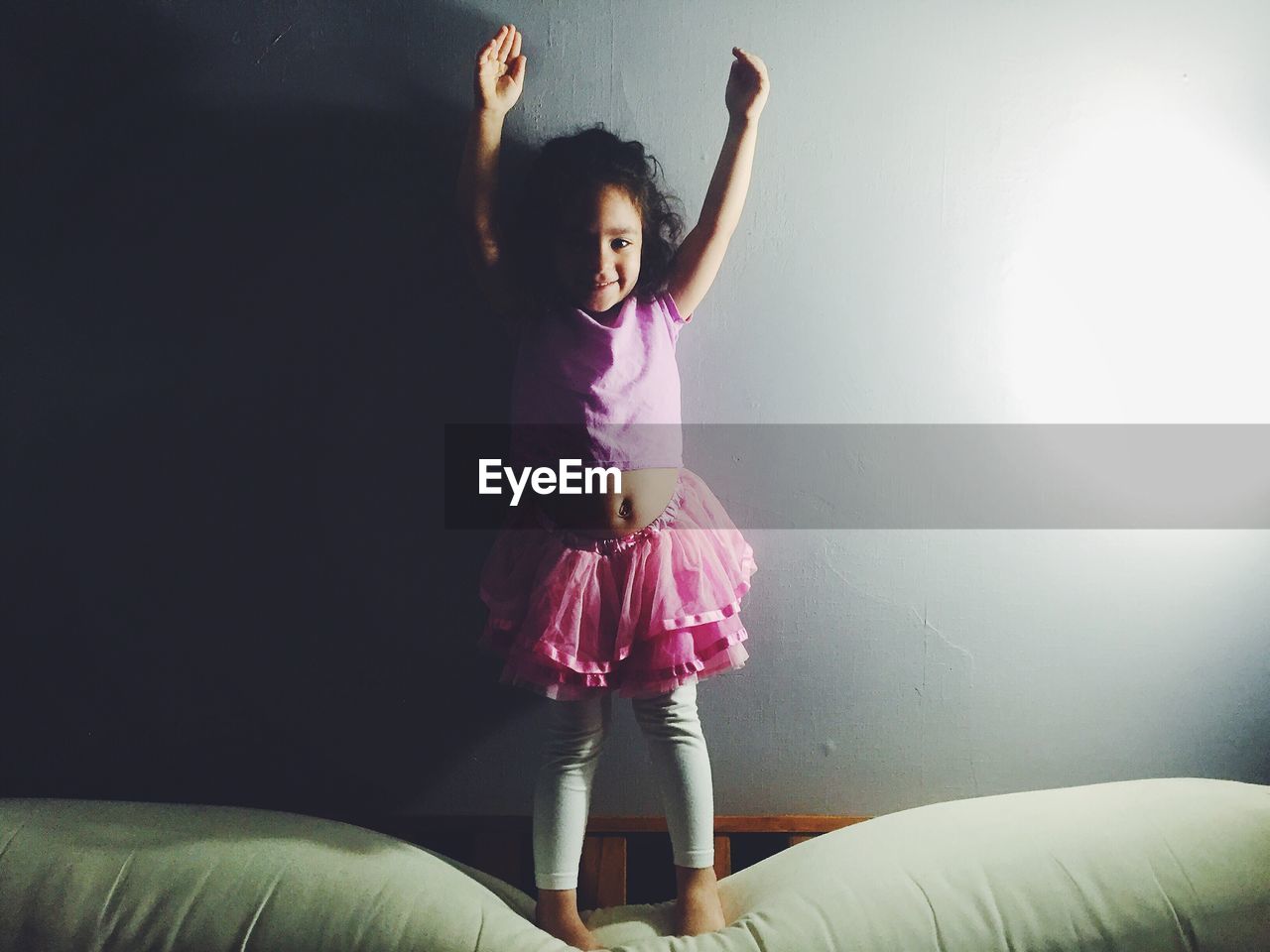 Portrait of happy ballerina standing on bed with arms raised