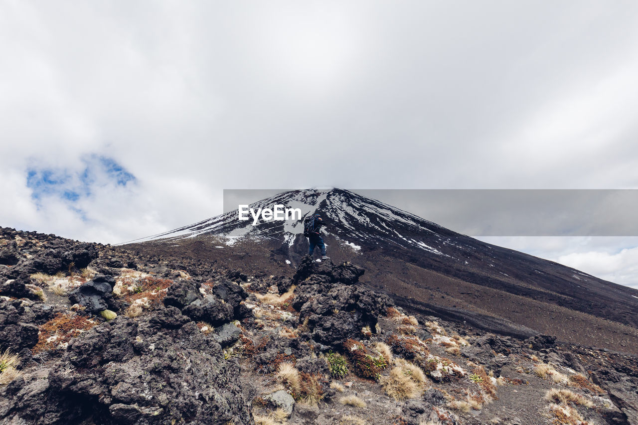 Mid distance view of hiker on rock at mountain against cloudy sky