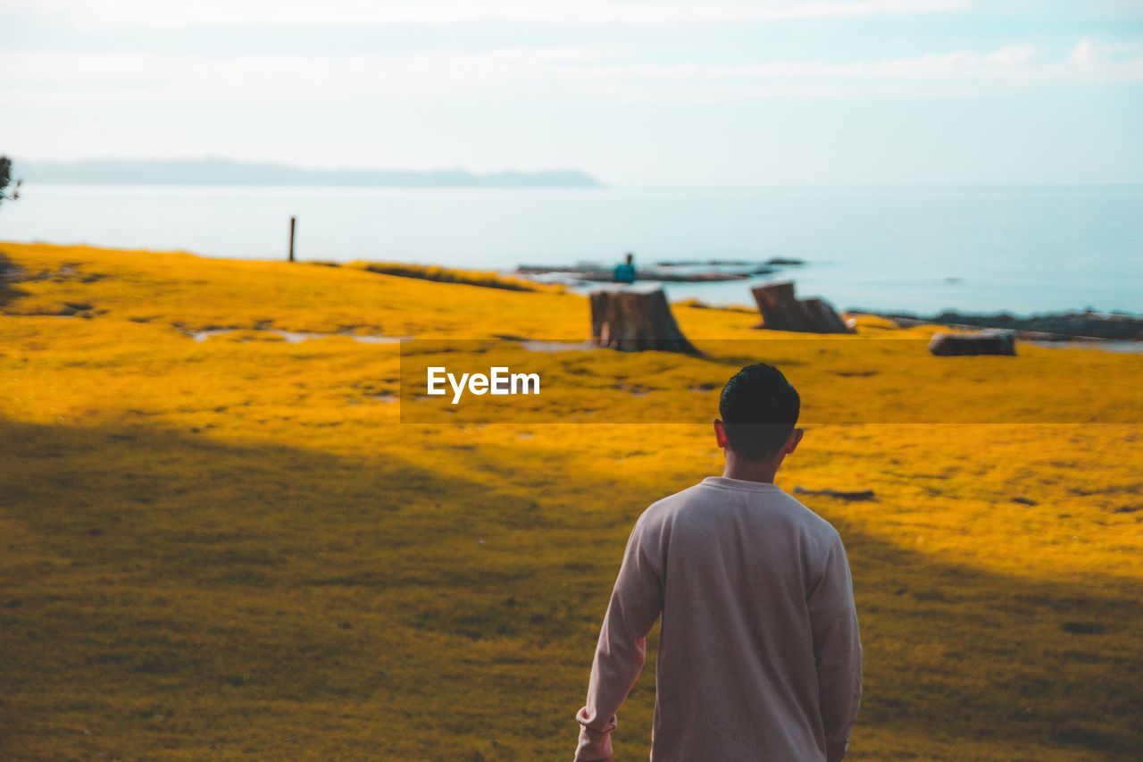 Rear view of young man walking on grassy field towards lake against sky