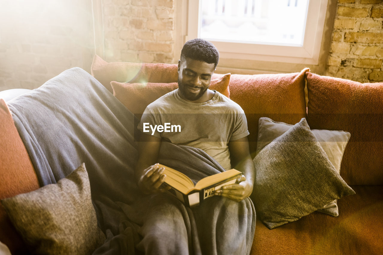 Smiling young man reading book while sitting on sofa with blanket at home