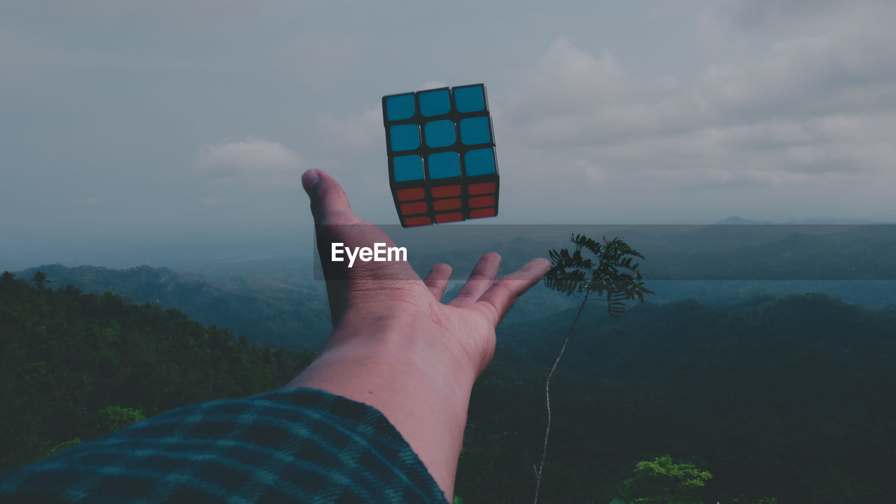 Cropped hand of woman reaching puzzle cube against sky