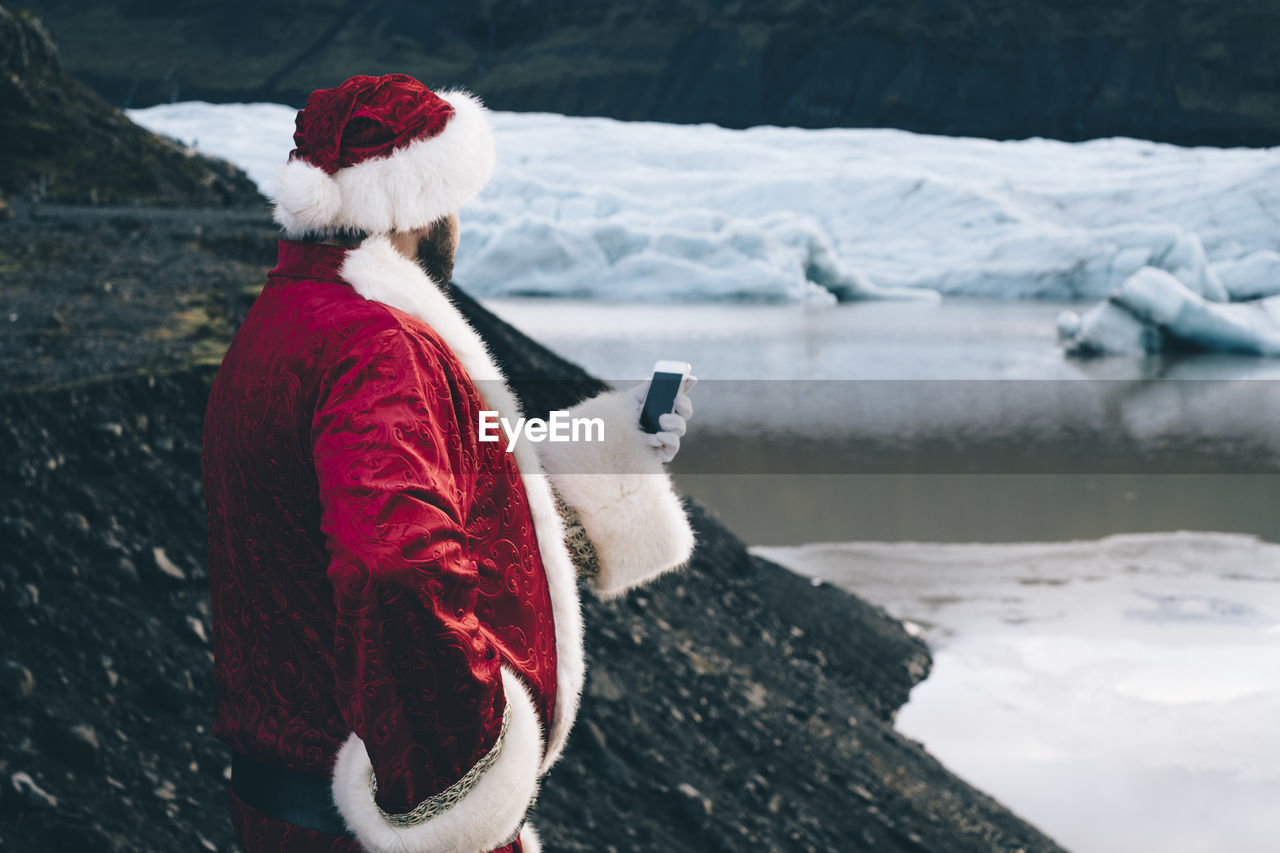 Side view of man wearing santa claus costume using phone by lake during winter