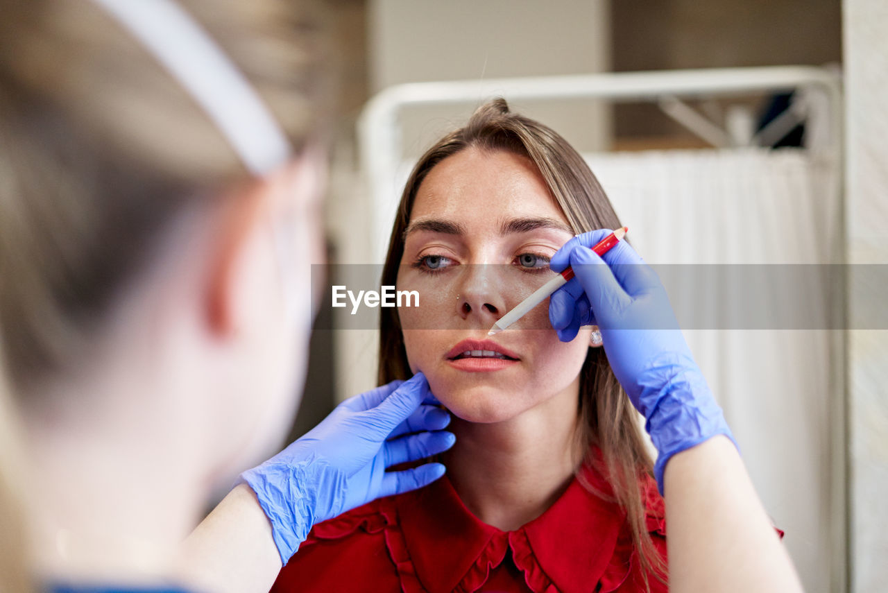 Blur beautician in latex gloves drawing line with white pencil while correcting shape of lips of young woman during work in aesthetic clinic