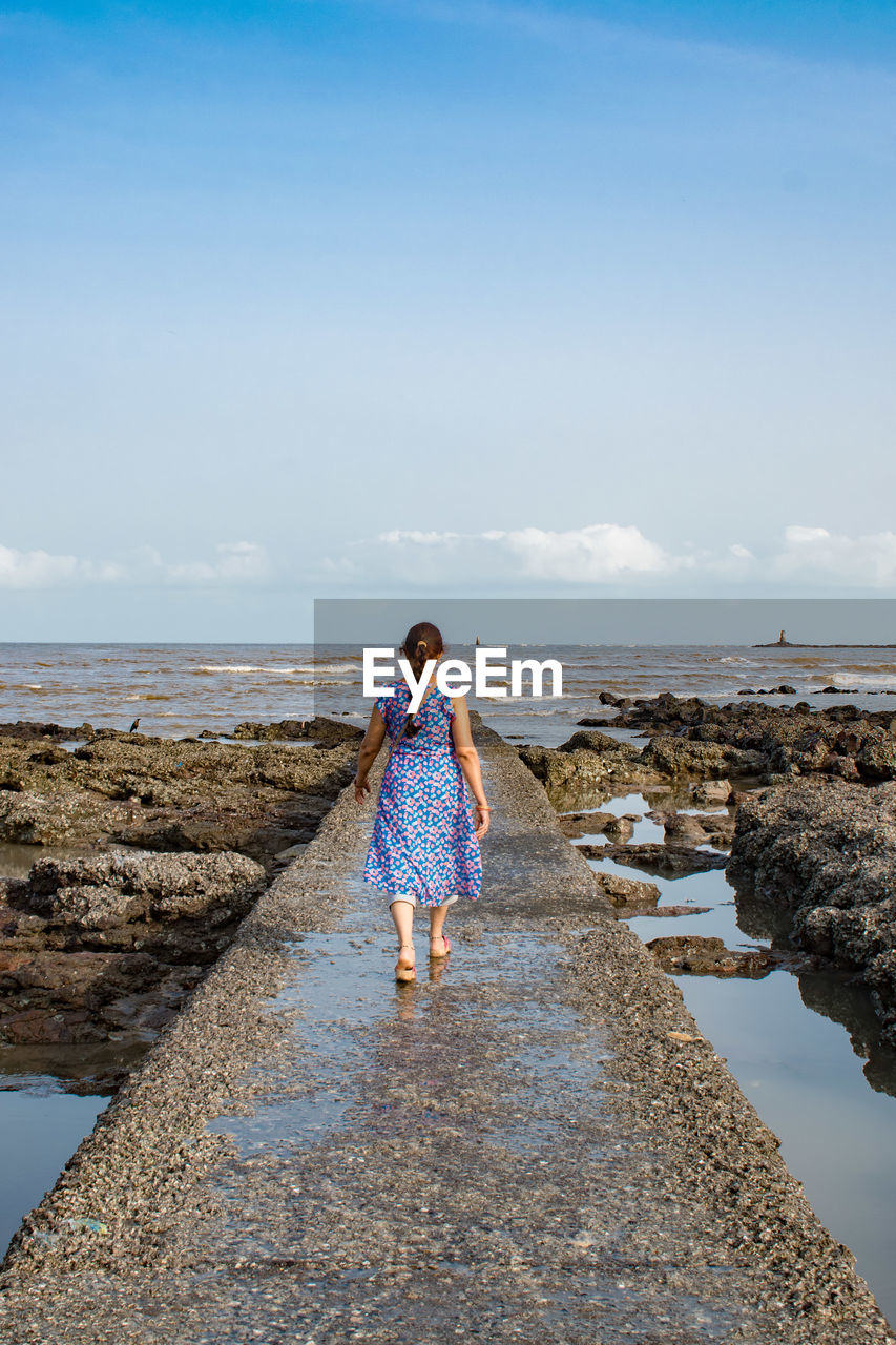 water, shore, sea, sky, one person, ocean, coast, vacation, full length, blue, beach, body of water, land, nature, rock, horizon, women, standing, leisure activity, sand, beauty in nature, horizon over water, day, casual clothing, rear view, wave, adult, lifestyles, holiday, tranquility, reflection, female, scenics - nature, trip, walking, child, cloud, outdoors, person, childhood, clothing, sunlight, dress, bay, tranquil scene, young adult, looking