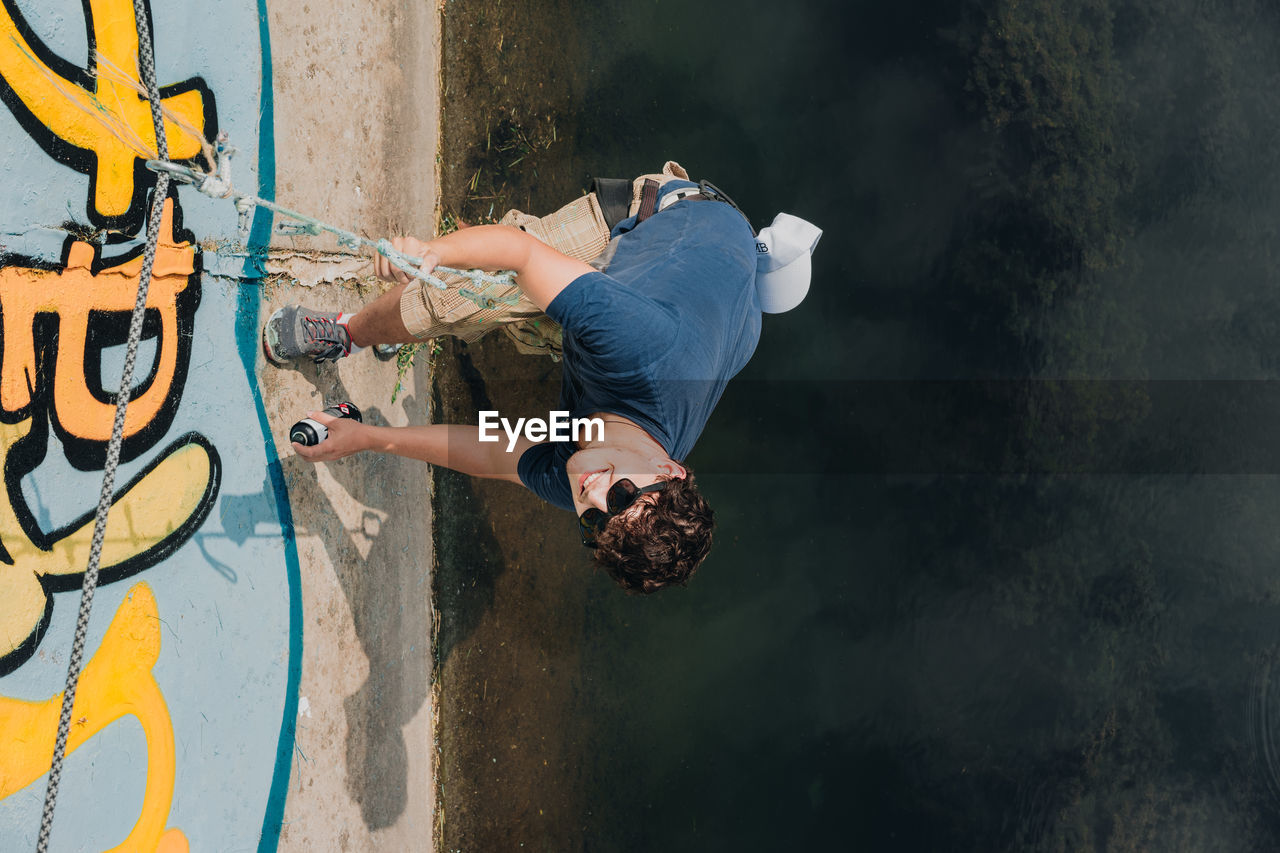 High angle portrait of man spray painting on wall over canal