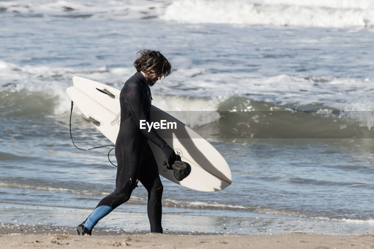 REAR VIEW OF MAN WITH SURFBOARD STANDING ON BEACH