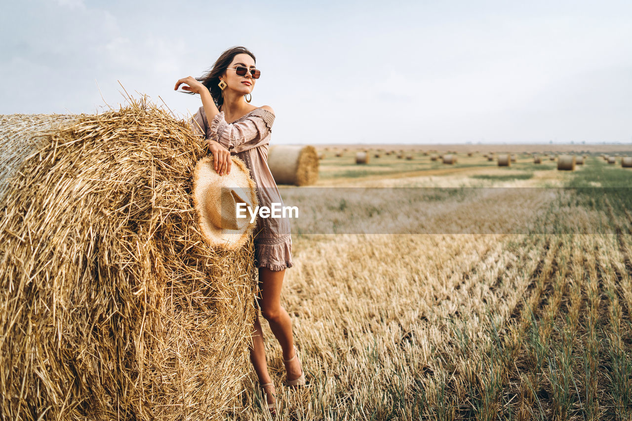 Smiling woman in sunglasses with bare shoulders on a background of wheat field and bales of hay.
