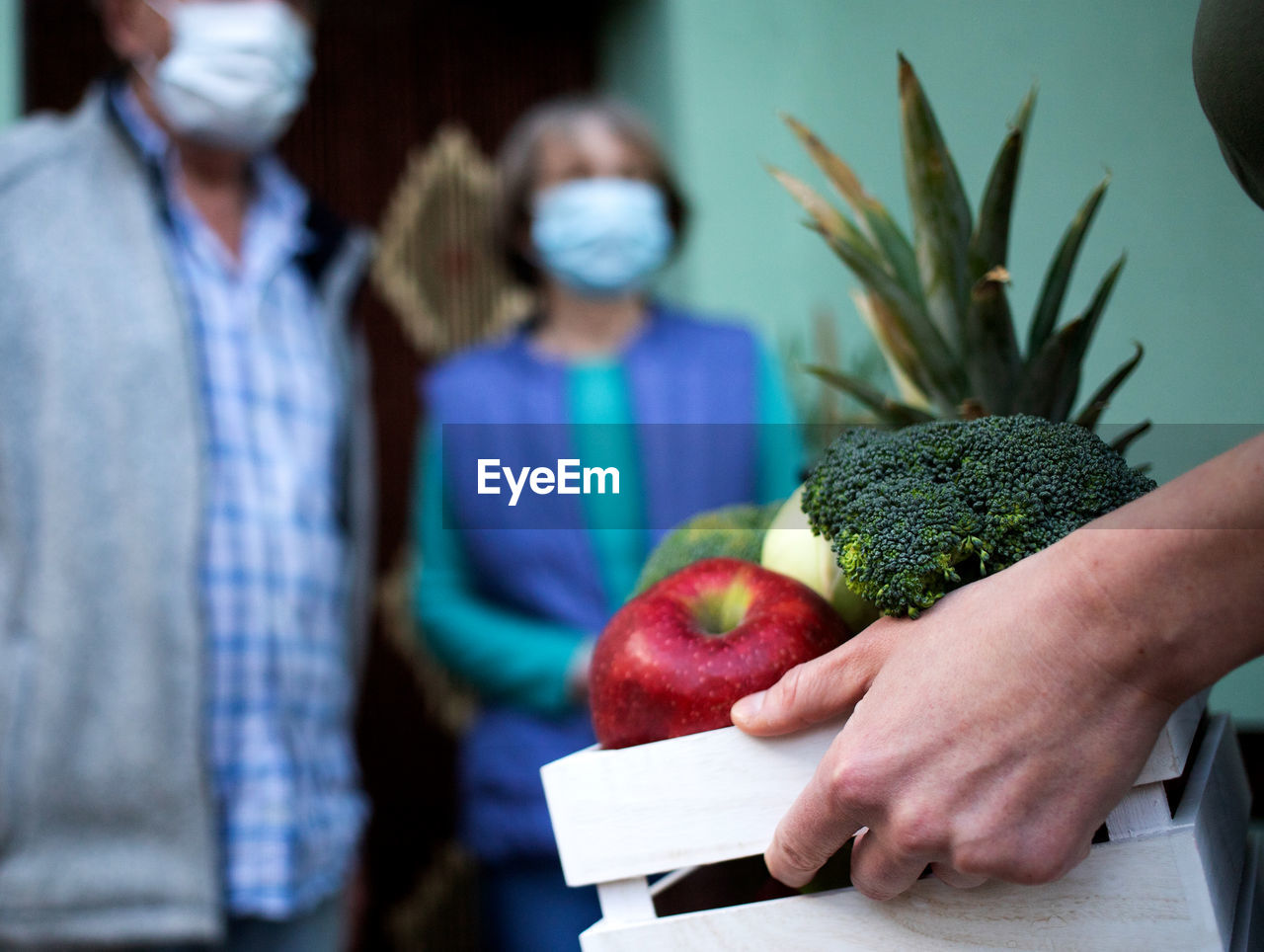 Hand of woman carrying crate with food for senior couple during pandemic