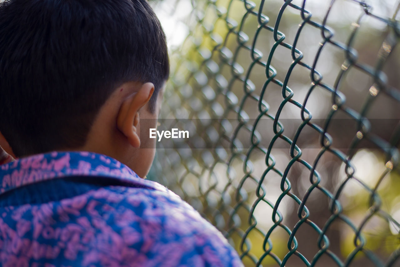 Rear view of boy looking through chainlink fence