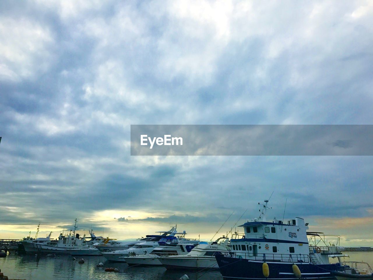 BOATS MOORED AT HARBOR AGAINST CLOUDY SKY