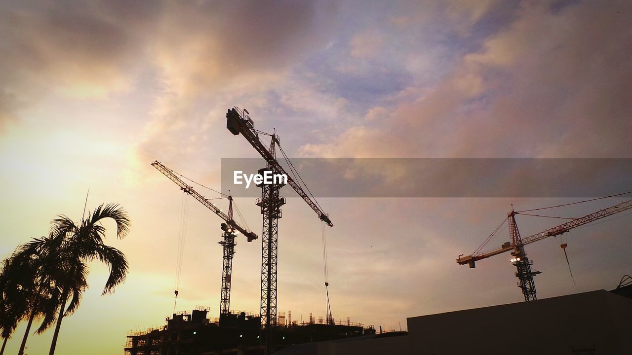 Low angle view of cranes against cloudy sky during sunset