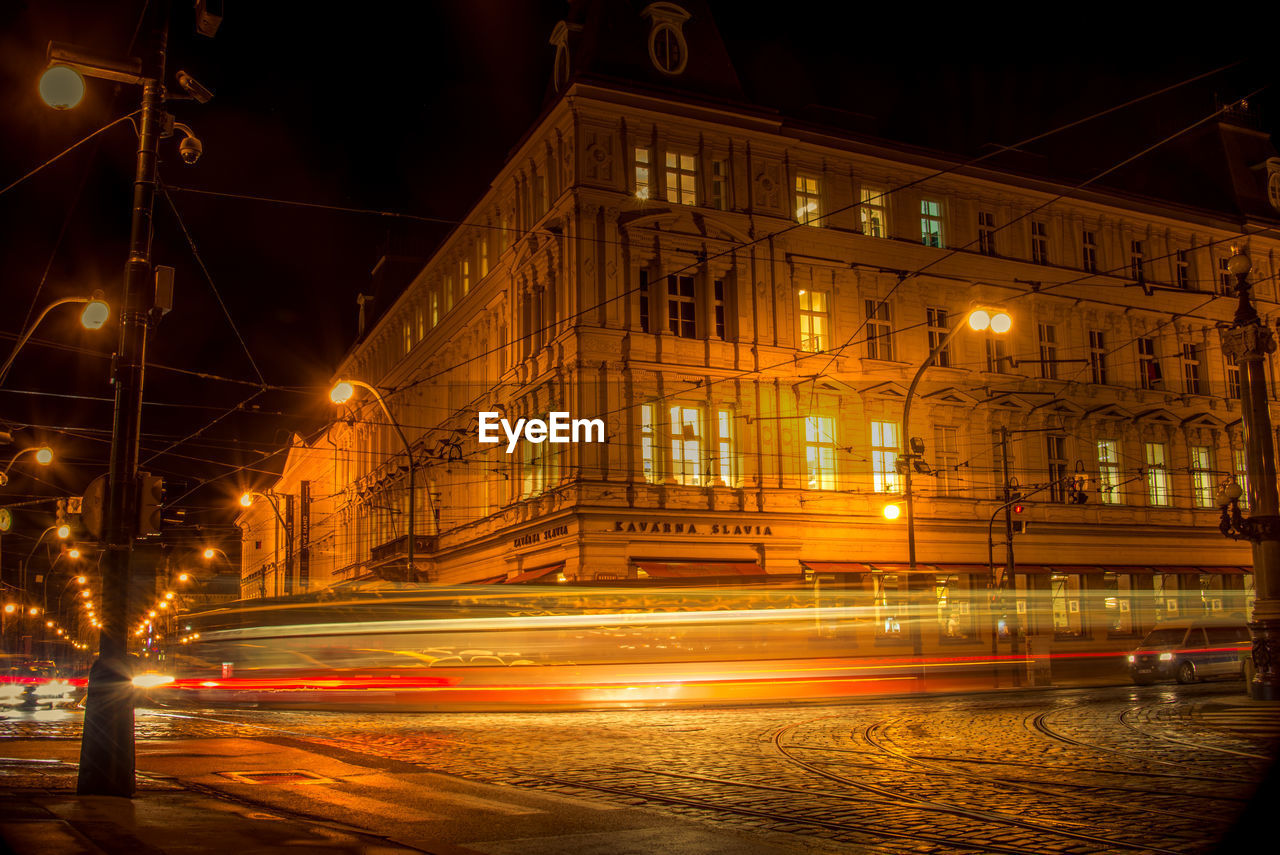 LIGHT TRAILS ON ROAD BY BUILDINGS AT NIGHT