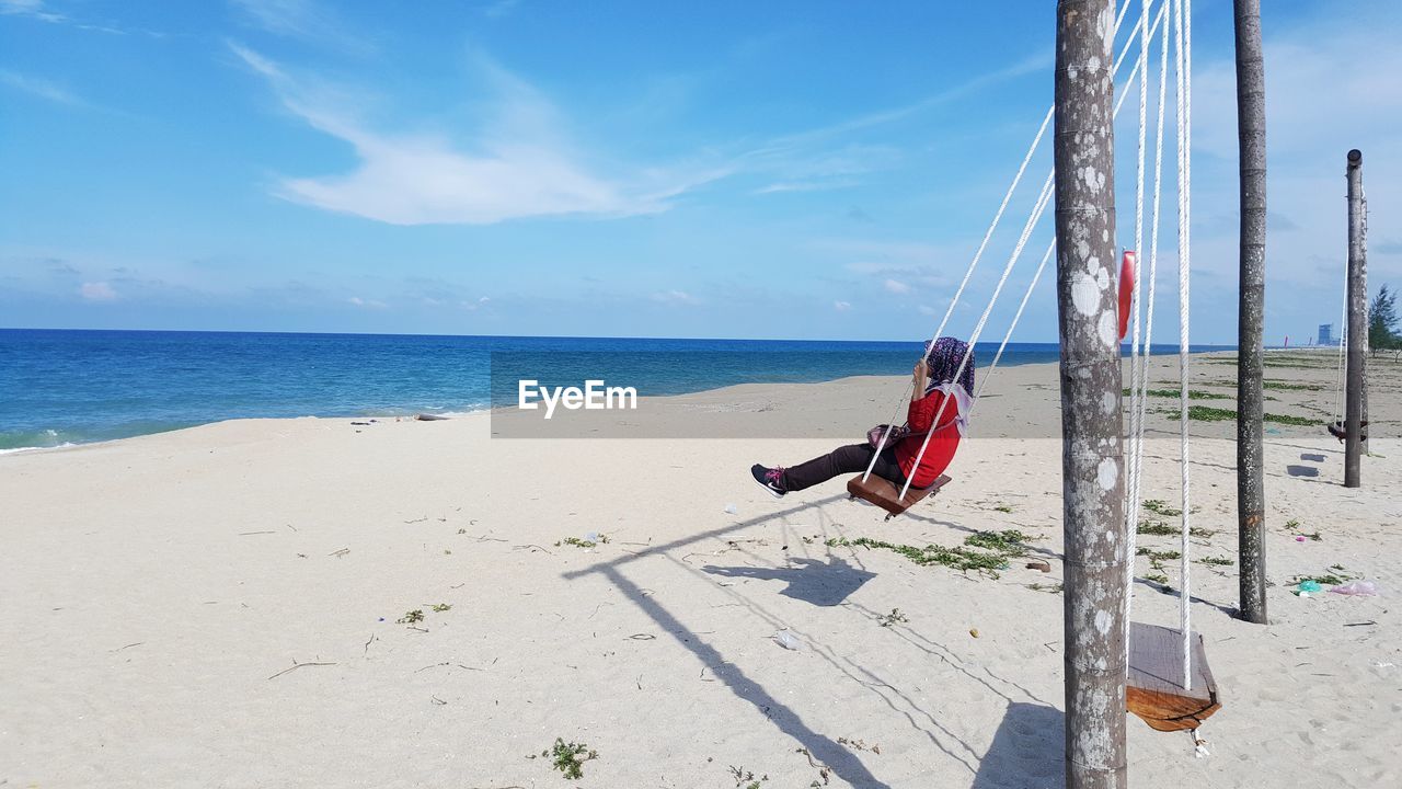 Woman on swing on beach against blue sky