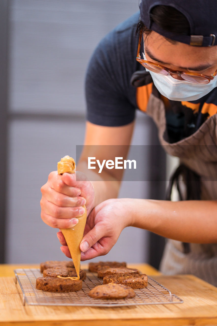 Focused asian man in medical mask holding piping bag with caramel sauce topping chocolate chip cookies on cooling rack