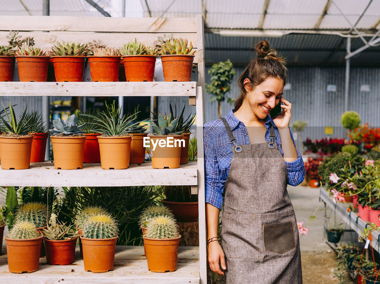 Happy lady in apron smiling and answering phone call while leaning on shelves with succulents during work in hothouse