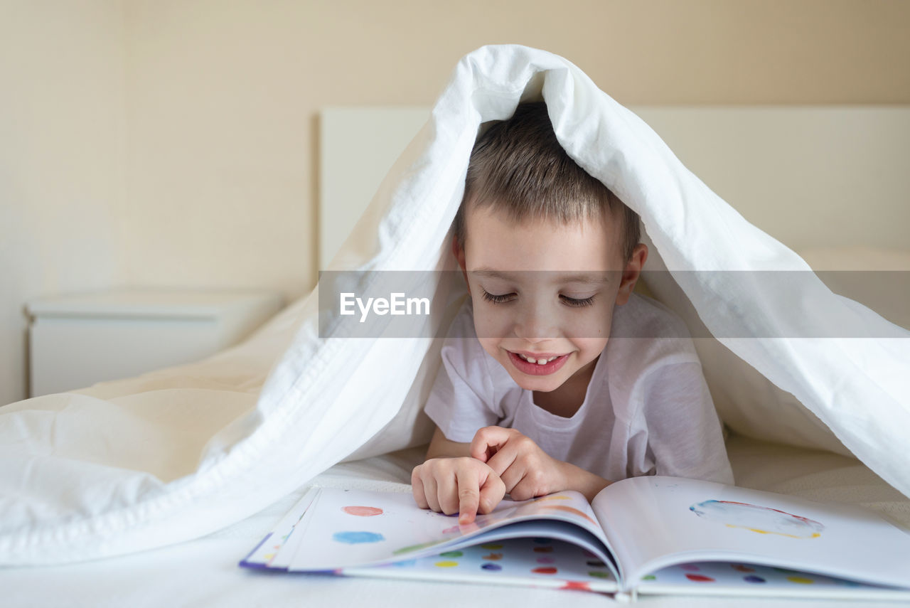 Adorable smiling kid lying down on bed under blanket, reading a book