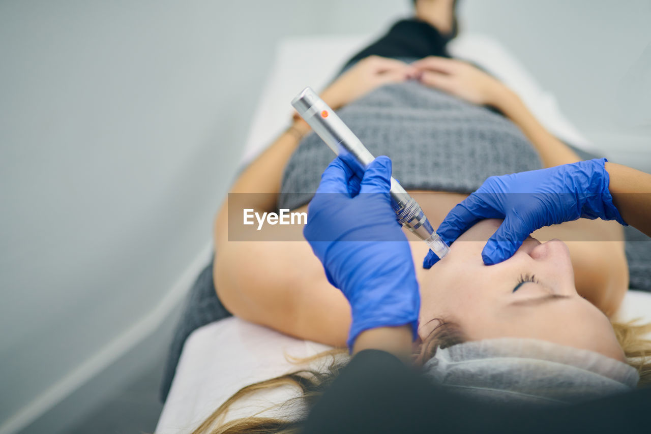 Cropped unrecognizable cosmetologist in uniform using derma pen with micro needles and cleaning face of client in beauty clinic