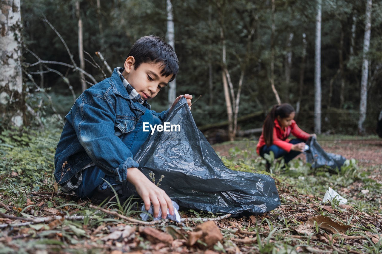 Ethnic volunteers with plastic bags picking rubbish from terrain against trees in summer woods in daylight