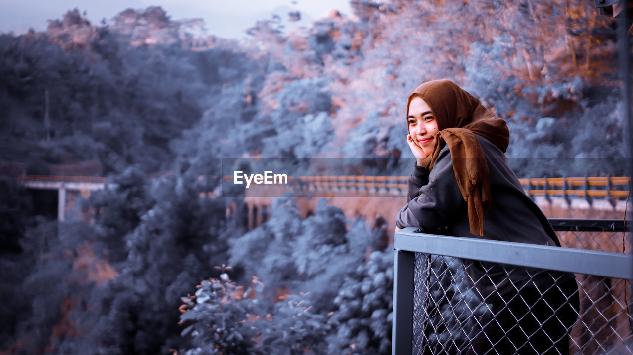 Smiling woman looking away while standing by railing during winter