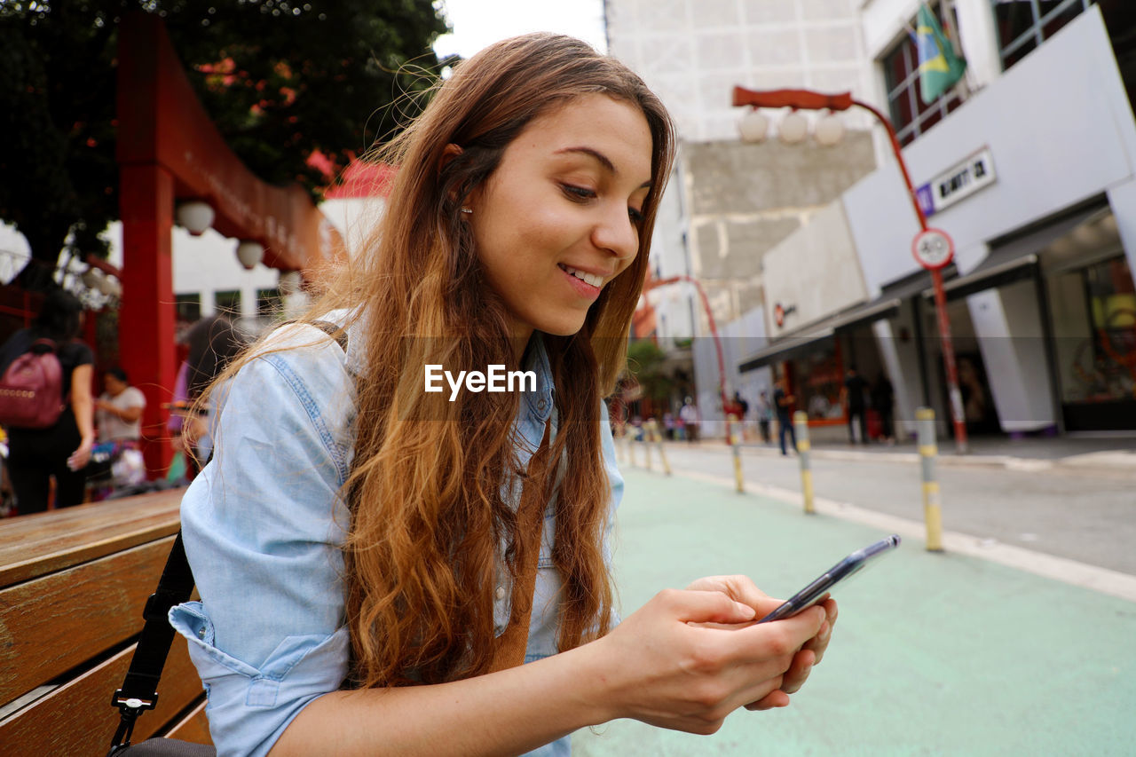 Smiling young woman using mobile phone against buildings in city