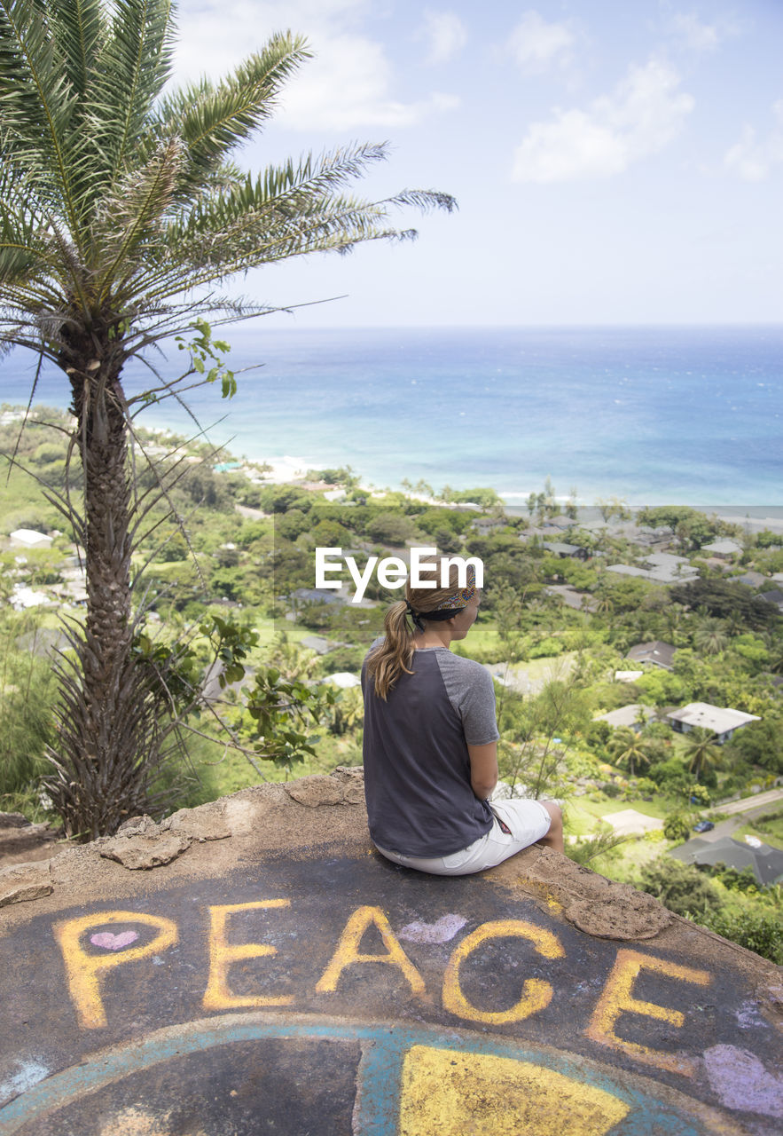 Rear view of woman looking at sea while sitting on cliff against sky