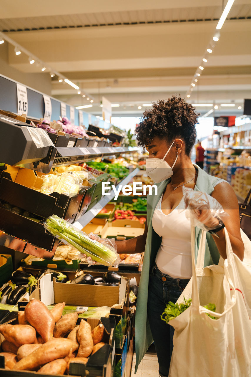 Side view of young african american female in protective mask and gloves choosing red apples while visiting grocery store during coronavirus pandemic