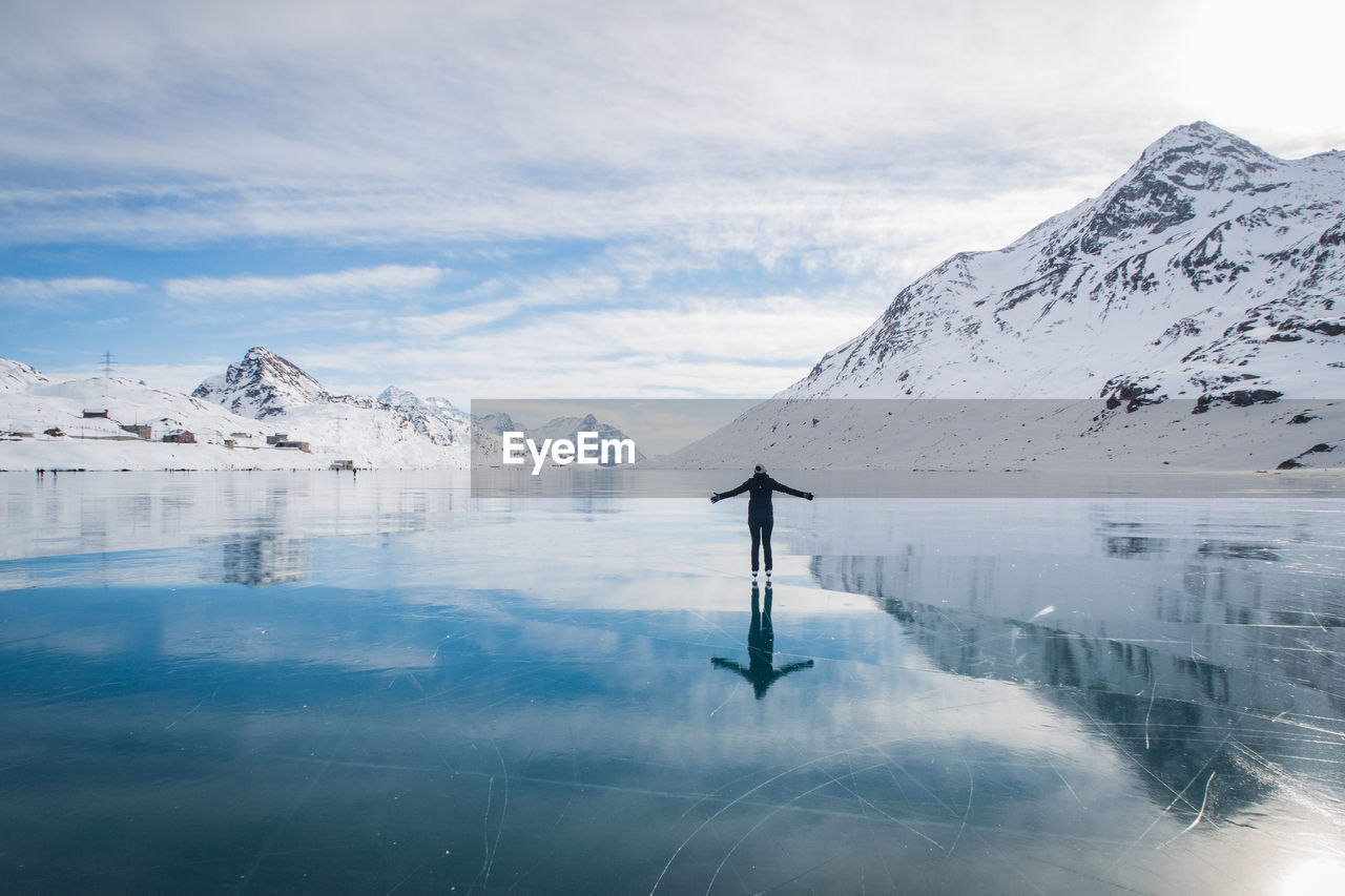 Woman standing on frozen lake by snowcapped mountain against cloudy sky